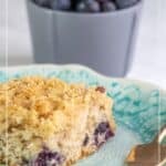 A slice of blueberry coffee cake with crumb topping sits on a blue and white plate, accompanied by a fork and a bowl of fresh blueberries in the background.