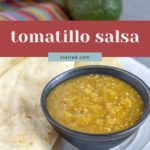 A bowl of vibrant tomatillo salsa sits on a plate next to a warm tortilla, with an avocado and a colorful cloth in the background.