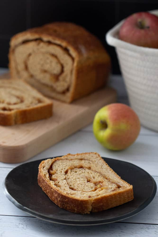 Apple cinnamon swirl bread slice on a black plate with loaf and apples in the background