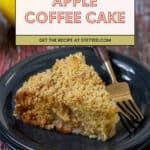 A slice of apple coffee cake with a crumb topping is served on a dark plate with a fork, highlighting the moist texture of the apple coffee cake. A lemon is blurred in the background.