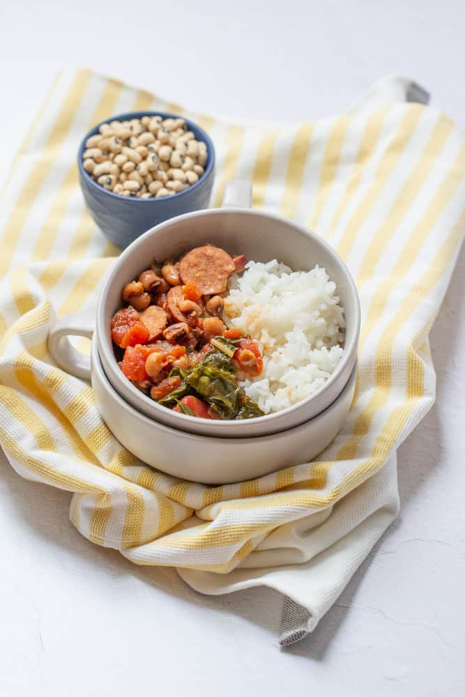 overhead of black eyed peas, sausage, tomatoes, greens, and rice in a stacked bowl on a yellow striped napkin