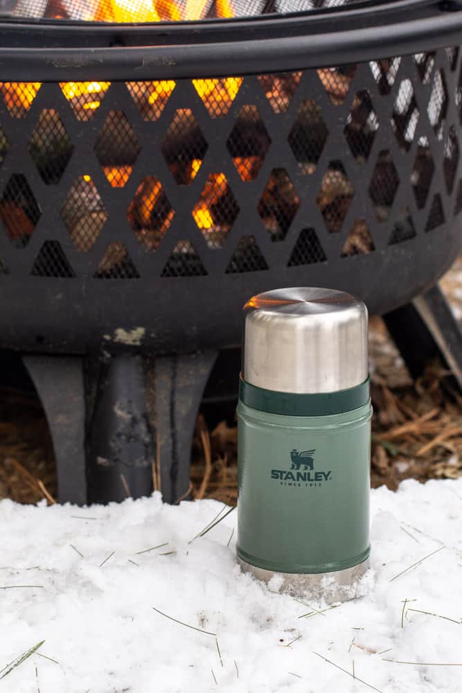 green Stanley food jar next to a fire pit in the snow