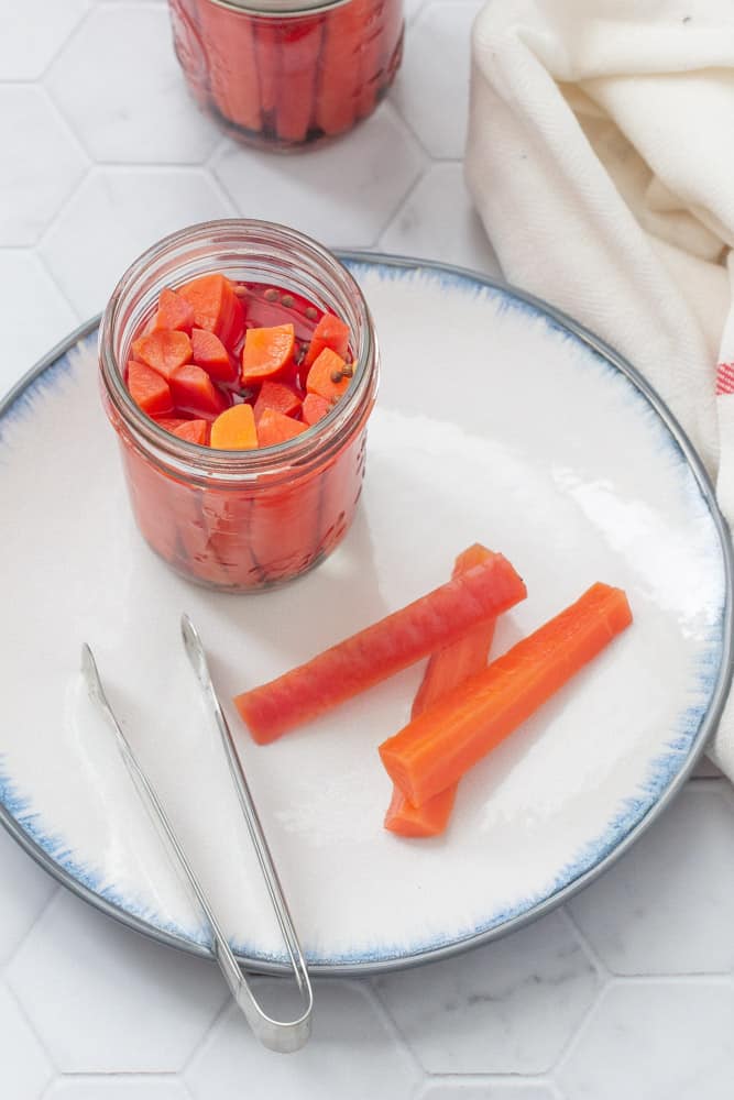 pickled carrots in a jar with carrots and tongs on a plate with a kitchen towel beside