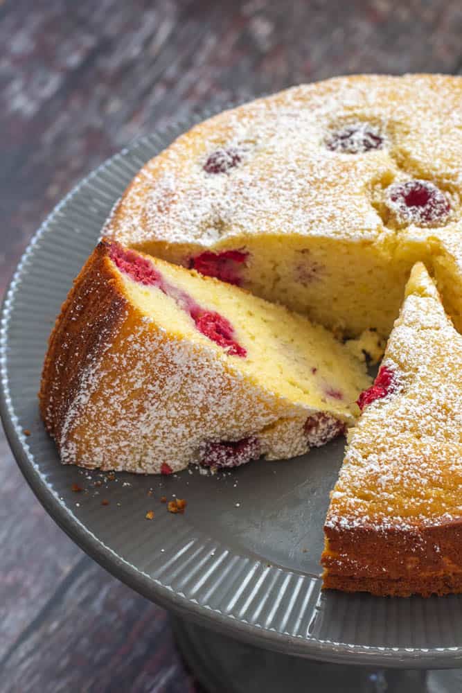 Close up of sliced ricotta cake with raspberries and lemon on a gray cake plate.