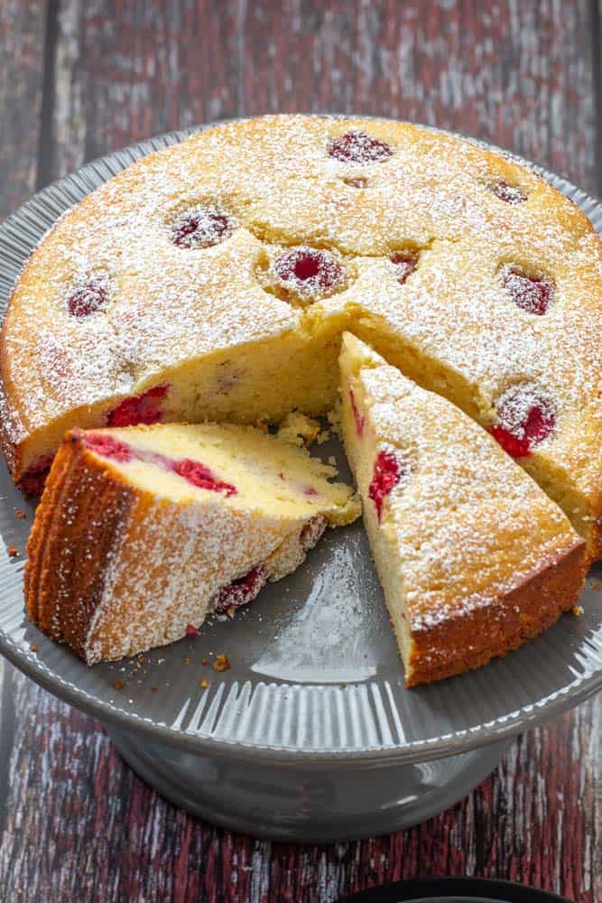 Sliced ricotta cake with raspberries and lemon on a gray cake plate.