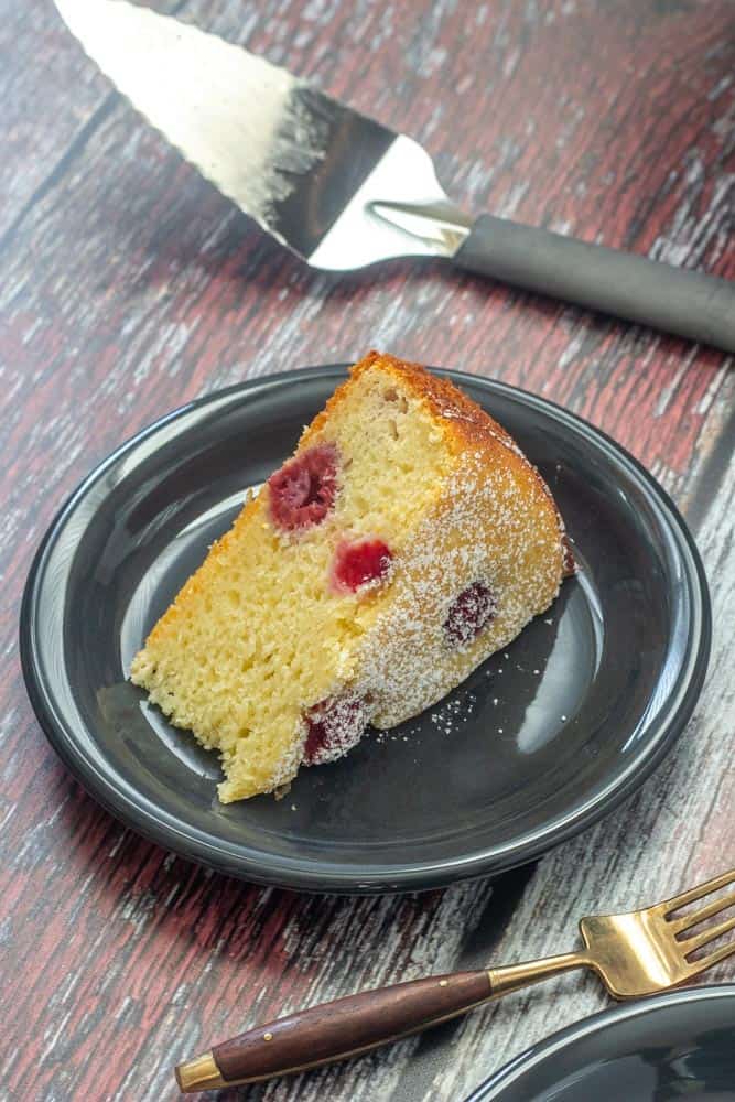 slice of ricotta cake with raspberries and lemon on a a plate with a fork and cake slicer