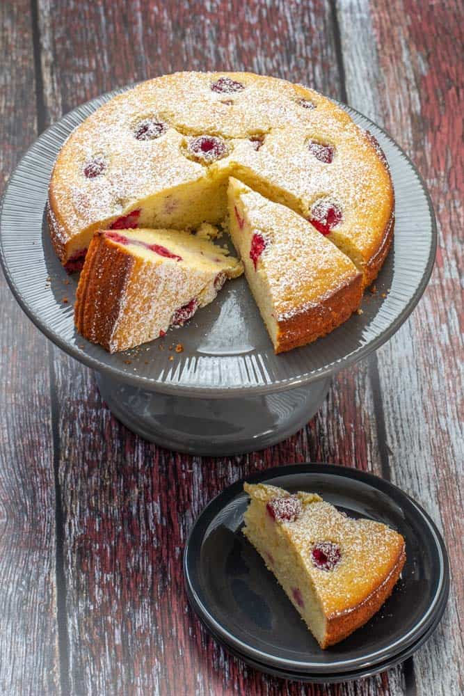 Sliced ricotta cake with raspberries and lemon on a gray cake plate with a slice off to the side on a black plate.