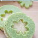 Green pistachio pudding cookies with a flower-shaped cutout, revealing a cream filling, are stacked neatly on a light wooden surface.