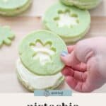 A hand holds a pistachio pudding cookie with a flower-shaped cutout, above stacked pistachio pudding cookies filled with white cream on a wooden surface.