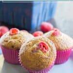 Three raspberry bran muffins in pink paper liners are displayed on a white plate, with whole raspberries and a blue container in the background, highlighting the fresh appeal of these wholesome raspberry bran muffins.