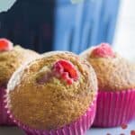 Three raspberry bran muffins in pink paper liners, each topped with a juicy raspberry, sit on a surface in front of a blurred background.
