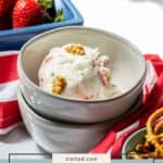 A bowl of strawberry pretzel ice cream sits on stacked white dishes, surrounded by fresh strawberries, crunchy pretzels, and a red-striped cloth nearby.