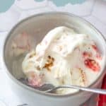 A bowl of strawberry pretzel ice cream with visible strawberry swirls and crunchy pretzel pieces, served with a spoon on a white surface.