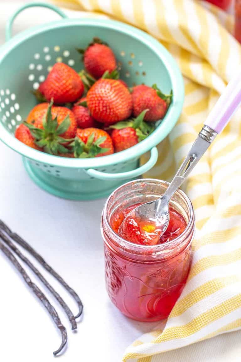 strawberry vanilla jam in a jar with strawberries, vanilla beans and yellow striped kitchen towel on a table