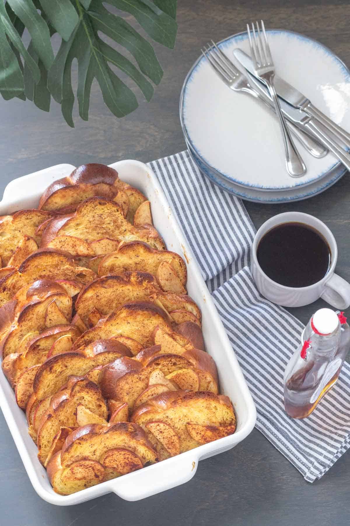 casserole dish of overnight apple cinnamon french toast with syrup and plates beside