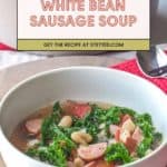 A bowl of hearty white bean sausage soup with chopped kale, sausage slices, tomatoes, and tender white beans sits next to a spoon and red napkin. A banner above reads "White Bean Sausage Soup.