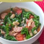 A bowl of hearty white bean sausage soup features kale, tomatoes, and sliced sausage, served on a wooden surface with a red napkin nearby.