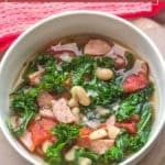 A bowl of hearty white bean sausage soup, filled with sliced sausage, white beans, tomatoes, and kale, rests on a wooden surface with a red cloth nearby.