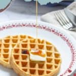 Two peanut butter waffles on a white plate with a pat of butter, being drizzled with syrup. A fork and knife rest in the background. Text at the top reads "PEANUT BUTTER WAFFLES.