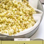 A metal bowl lined with parchment paper filled with flavorful pesto popcorn, the kernels coated in vibrant green seasoning and labeled "pesto popcorn.