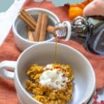 A bowl of pumpkin baked oatmeal topped with whipped cream, as syrup is poured on top; cinnamon sticks and pumpkin pieces in the background create a cozy fall vibe.