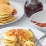 A plate of bacon pancakes with a fork, crispy pieces of bacon, and a stack of fluffy bacon pancakes in the background, accompanied by a pitcher of syrup.