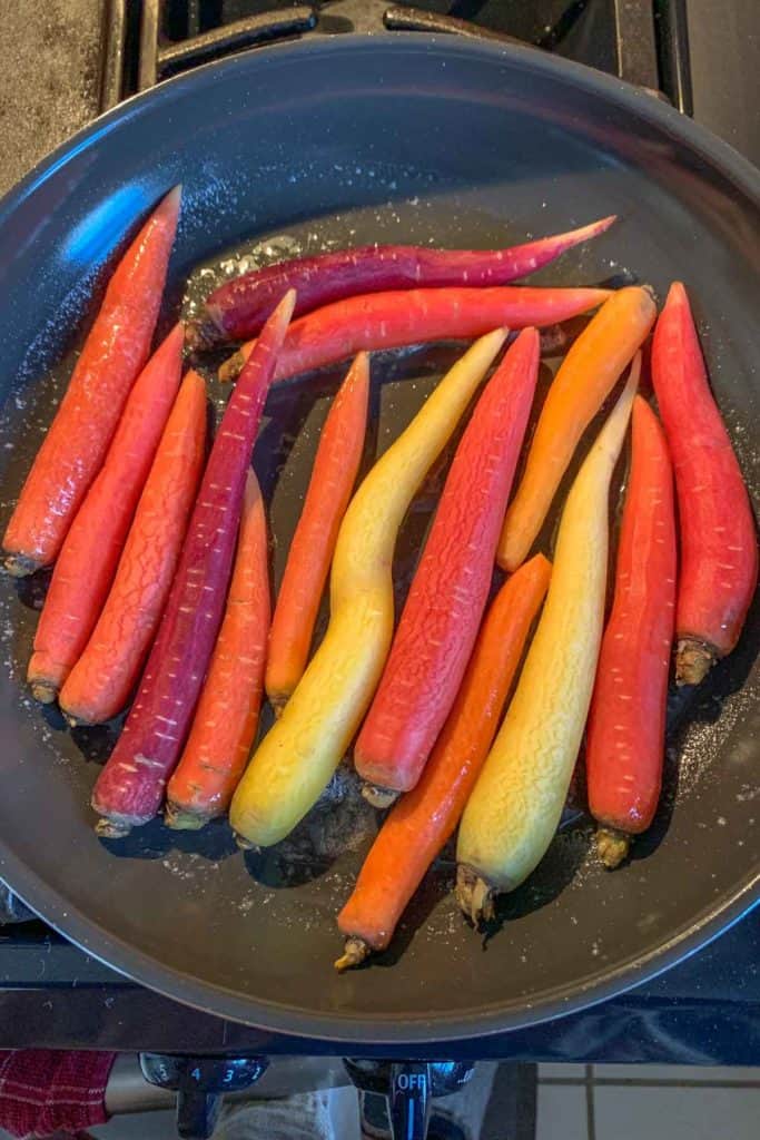A frying pan on a stovetop with multicolored carrots, including red, orange, and yellow varieties, being sautéed for delicious maple glazed carrots.