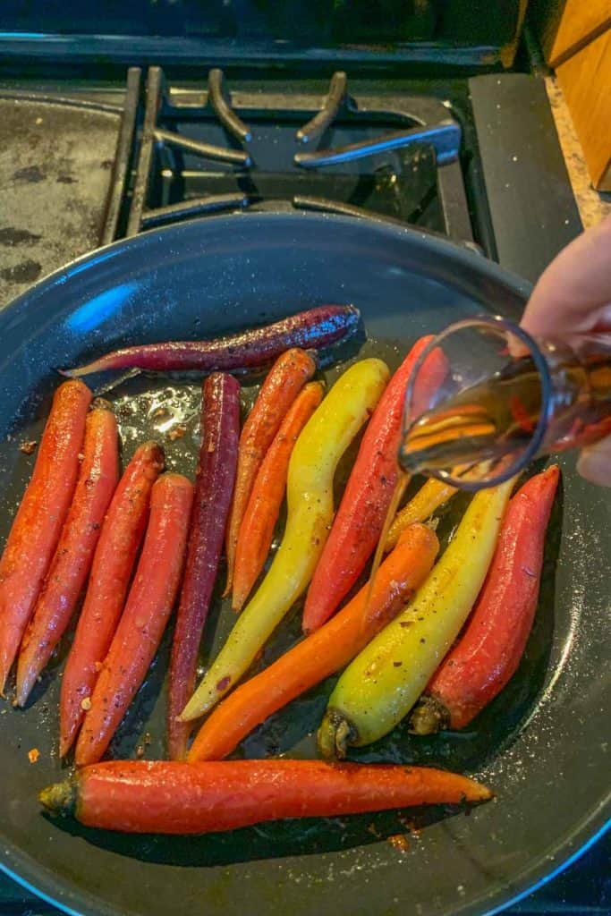 A hand pours liquid over assorted colorful carrots in a black pan on a stovetop, creating delicious maple glazed carrots.
