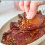 A hand lifts a slice of million dollar bacon, glazed and sprinkled with red pepper flakes, from a dish filled with similar savory slices; text above reads "million dollar bacon.