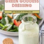 A glass jar of green goddess dressing recipe sits in front of a salad bowl with greens and strawberries, next to a cork and a striped cloth.