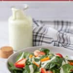 A bowl of spinach salad with sliced strawberries and carrots drizzled with green dressing, next to a bottle of green goddess dressing—perfect for trying your own green goddess dressing recipe at home.