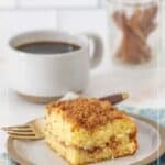 A slice of cinnamon coffee cake rests on a plate with a fork, accompanied by a cup of black coffee and a jar of cinnamon sticks in the background.