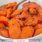 A white bowl filled with honey roasted carrots, sliced and garnished with herbs, sits invitingly with a metal spoon in the background.