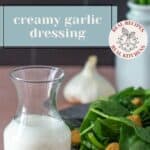 A glass jar of creamy garlic dressing sits next to a plate of fresh spinach and croutons, with a garlic bulb and vase of chives in the background, highlighting this flavorful creamy garlic dressing.