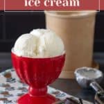 A red bowl filled with creamy vanilla bean ice cream sits on a patterned cloth, with an ice cream scoop, whole vanilla beans, and a tub of vanilla bean ice cream in the background.
