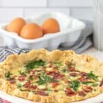 A freshly baked bacon spinach quiche in a red pie dish sits on the counter, with a carton of eggs and a kitchen towel in the background.