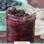 A glass jar filled with homemade blueberry pie filling sits on a table, with a spoon in the jar and a bowl of fresh blueberries in the background.