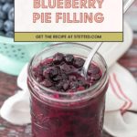 A mason jar filled with homemade blueberry pie filling sits on a wooden surface, with a spoon inside and a bowl of fresh blueberries in the background.
