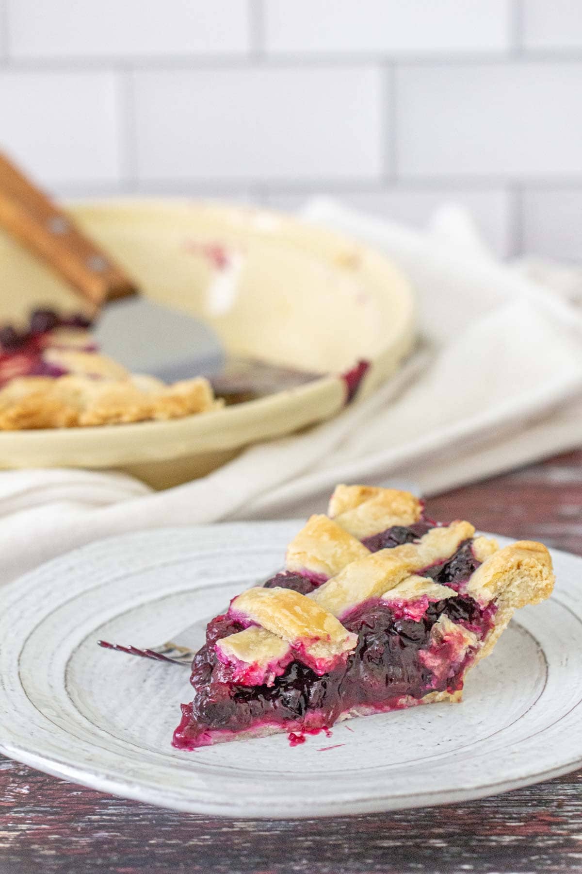 A slice of lattice-topped pie with blueberry pie filling sits on a plate, while the rest of the pie and a pie server can be seen in the background.