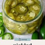 An open glass jar filled with pickled jalapeños in brine, surrounded by whole and sliced fresh jalapeños on a light surface.