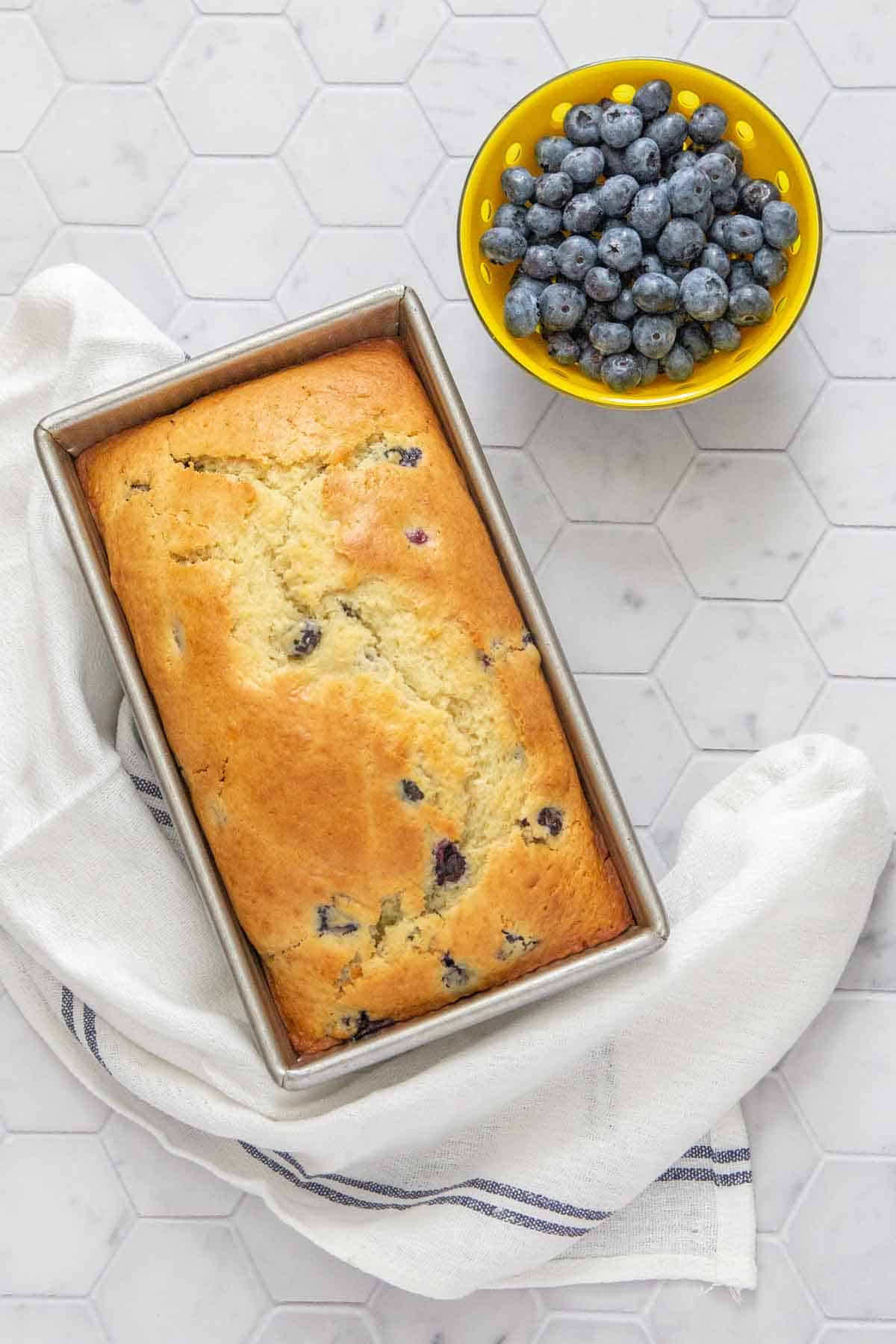 loaf of blueberry bread in baking pan with bowl of blueberries