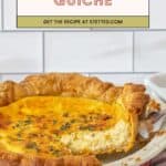A delicious cheese quiche with a slice being served from a red ceramic dish, topped with fresh herbs, sits on a striped towel against a white tiled background.
