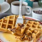 A fork holds a bite of chocolate chip waffles over a plate with syrup, with coffee and extra chocolate chips in the background. Text reads "Chocolate Chip Waffles" and "stetted.com".