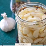 A jar of pickled garlic sits on a table, accompanied by a head of garlic and a jar of spices in the background.