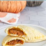 A plate with pumpkin empanadas, one cut in half to reveal its spiced filling, with another whole empanada and a festive pumpkin decoration in the background.