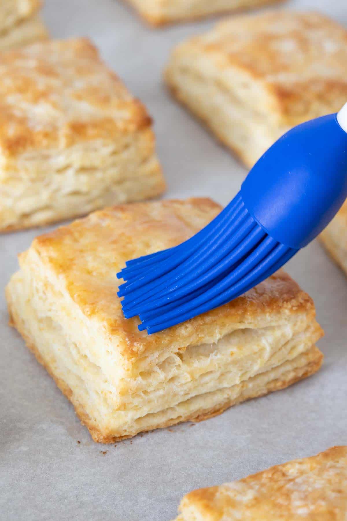 Blue pastry brush brushing melted butter onto buttermilk biscuit.