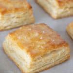 Close up of buttermilk biscuits on a baking sheet with melted butter and flaky salt on top of the biscuit in center.