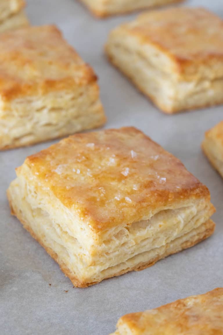 Close up of buttermilk biscuits on a baking sheet with melted butter and flaky salt on top of the biscuit in center.