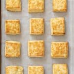 Twelve square buttermilk biscuits are neatly arranged in rows on a parchment-lined baking sheet, viewed from above.