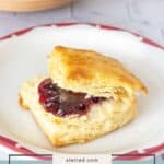 A flaky buttermilk biscuit split in half with a layer of dark red jam sits on a white plate with a red rim, making classic buttermilk biscuits even more irresistible.